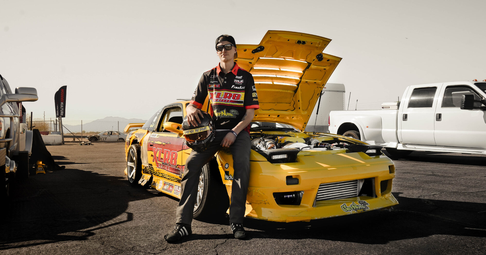 Tyler Krenek leaning on the hood-up XLR8 Nissan S13 in the paddock, helmet in hand